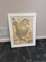 Full view of framed vintage-style North America map leaning against white wall on dark carpeted floor.