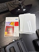 Box and loose stack of HP Premium Plus glossy photo paper sheets on black desk with keyboard and monitor stand visible.