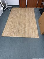 Natural jute rug laid out on a blue carpeted floor, showing its full rectangular shape and woven texture.