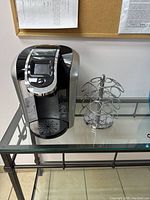 Front view of black and silver Keurig coffee machine with water drip tray and pod holder beside it on glass table.