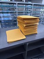 Stack of large yellow kraft paper envelopes, both plain and bubble-lined, on a worktable showing that there are many envelopes in this lot.