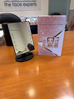 Photo of a black light-up makeup mirror on a wooden table, with its pink product box featuring images of the mirror and usage instructions.