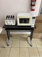Full view of the Sharp microwave and T-fal toaster on small black table against a white wall with fire extinguisher in background.