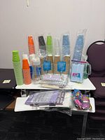 Photo of multiple stacks of disposable paper cups in green, orange, black, blue, white, and kraft brown, along with packs of straws and packaged tablecloths and balloons.