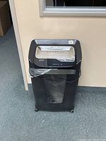 Front view of black paper shredder showing top shredding slot, control panel, and transparent bin at bottom with caster wheels.