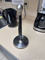 Paper towel holder on kitchen counter with between a black electric kettle and coffee maker in the background.