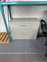 Front view of closed small grey metal filing cabinet with 2 drawers, placed on the floor beside a teal metal shelving unit and office chair.