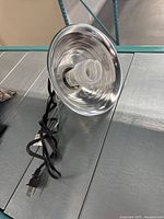 Silver metal reflector spotlight with a spiral compact fluorescent bulb and attached black power cord on a gray surface.