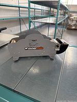 Front side view of the gray metal wet paper sealer with Sticker Mule logo, sitting on metal surface with shelving in background.