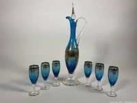 Full set view showing one tall blue glass decanter with clear handle and stopper and six matching stemmed glasses, all with gold gild decoration.