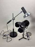 Two black lamps side by side, one spotlight style with barn doors on tripod base, one cantilever adjustable arm desk lamp with weighted round base