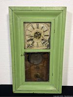 Front view of antique pendulum wall clock with pale green wooden frame, worn Roman numeral dial and glass door.