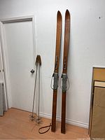Full view of the pair of wooden skis and two wooden ski poles leaning against a white wall.