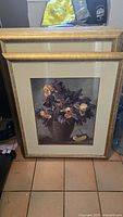 Full view of flower and fruit picture with gold frame resting against a wall on a tiled floor.