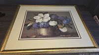 Framed flower picture showing white and purple flowers in a gold textured frame on a dark wood surface.