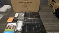 Photo showing the solar street light components laid out with solar panel, LED unit, mounting hardware, remote control, and user manuals, with product box in background.