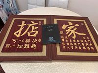 Photo of two wooden plaques side by side on table. Right plaque with single large character and multiple vertical gold text lines. Left plaque with large gold character and two rows of gold script. Red background, gold border, wood frame.