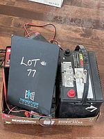 View of assortment showing automotive battery, black battery charger casing labeled 'LOT 77', multiple cables and connectors in box.