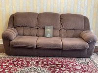 Front view of brown three-seat couch showing textured fabric upholstery, three seat cushions, and puffy padded arms and backrest under natural lighting on patterned red rug.