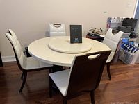 Round dining table with visible lazy Susan and 5 cream upholstered chairs around it. Room setting with items in background.