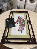 Framed embroidery with floral still life, shown upright on a round table against dining chairs and boxed items in the background.