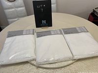 Three unopened sets of white IKEA Alvine Spets curtains on a round table with a black lot number sign