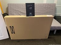 Photo of large unopened box showing IKEA branding and indicating it contains the desk, placed on floor against a wall.
