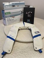 Front view of the PLCME-N01 Itap shoulder massager in white and blue, displayed in front of the original product box and a black sign with lot number