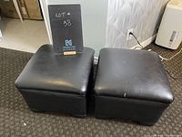 Two black faux leather ottomans placed side by side on carpet showing top and front surfaces with some wear and indentations.