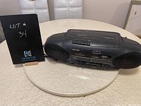 Black Sharp portable radio with built-in speakers, CD player on top, cassette player in front, pictured on white circular table.