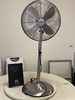 Front view of Noma upright extendable fan on a round table, chrome finish with visible wear on base.