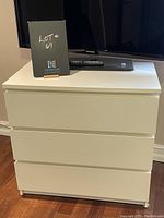 Front view of white three drawer dresser with TV placed on top and lot number card in front.