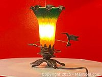 Front side view of the lamp showing the flower-shaped glass shade and a hummingbird figure on a metal rod.