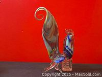 Front view of vintage Murano glass rooster with multicolored tail and broken leg on red background.