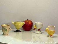 Four vintage hand-painted egg cups displayed next to an apple for size reference.