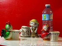 Overview showing green rooster, white watering can, small human-like figure with yellow hat, and red bird figurine with a water bottle for scale