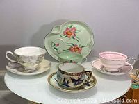 Photo showing three sets of teacups and saucers on a white table including green floral design, pink and white cups.