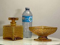 Amber diamond pattern square glass bottle with stopper and carnival glass pedestal candy dish with floral pattern placed side by side with water bottle for scale.