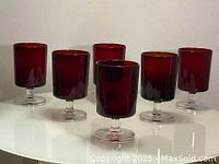 Six red ruby wine glasses on clear stems and bases displayed on a white surface against a plain light background, showing front and side views.