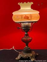 Full view of the vintage hurricane lamp showing the lamp with painted glass shade and ornate solid brass base on black surface against red wall.