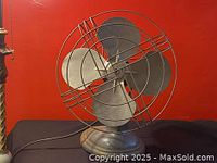 Front view of the vintage industrial metal desk fan showing full fan and base against a red background.
