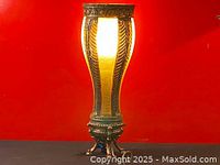 Front view of the lamp lit, showing fiberglass shade with decorative leaf patterns and ornate solid brass base.