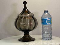 Overall front view of the olive grey glass candy jar with lid, next to a water bottle for scale.
