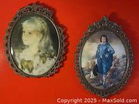 Pair of ornate brass Victorian style oval picture frames hanging on a red wall, one containing a portrait of a young girl and the other a portrait of a boy in a blue outfit.