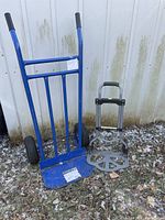 Image of two hand carts standing outdoors on grass and leaves. Larger blue metal cart on left and smaller silver folding cart on right.