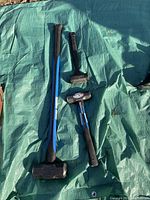 Four assorted sledge hammers laid on a green tarp, showing the full length and design of each hammer including black rubberized handles with blue accents and metal heads.