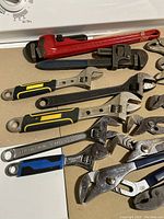 Wide view of assorted wrenches including large red pipe wrenches, adjustable wrenches with black, yellow, and silver handles, and locking pliers arranged on a beige surface.
