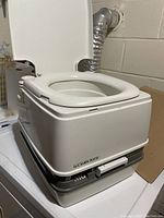 Side view of the Le Campa Potti MG portable toilet with lid open showing the toilet seat and some supplies.