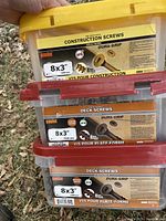 Three stacked containers of 8x3 inch construction and deck screws with clear plastic boxes, red and yellow lids, white handles. Top container yellow lid labeled for construction screws, bottom two containers with red lids labeled deck screws.