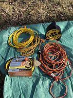 Six heavy duty extension cords laid out on a green tarp, three yellow and three orange including one coiled on a black and yellow multi-outlet reel.